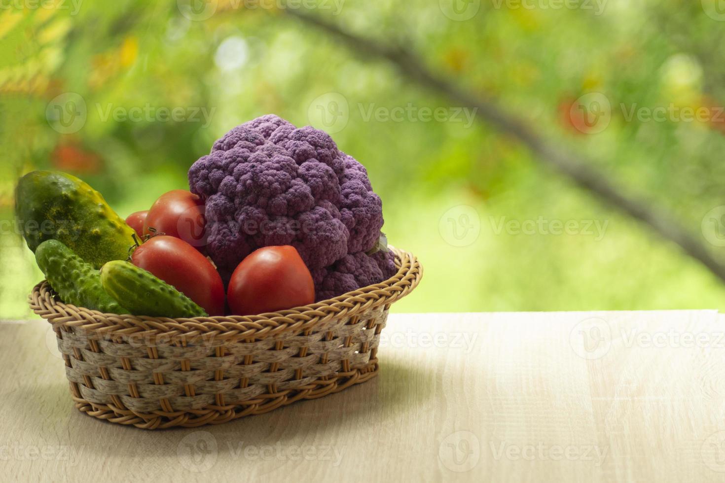 Fresh ripe purple cauliflower, red tomatoes and green cucumber in basket. Healthy food on table on defocus autumn background. photo