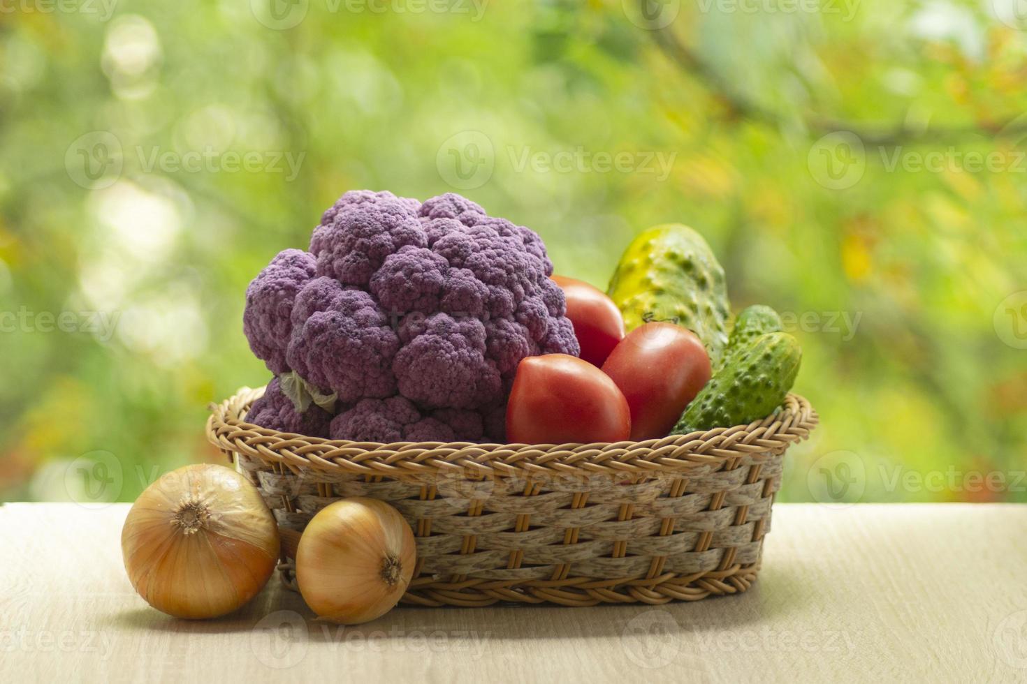 Fresh ripe purple cauliflower, onion, red tomatoes and green cucumber in basket. Healthy food on table on defocus autumn background. photo