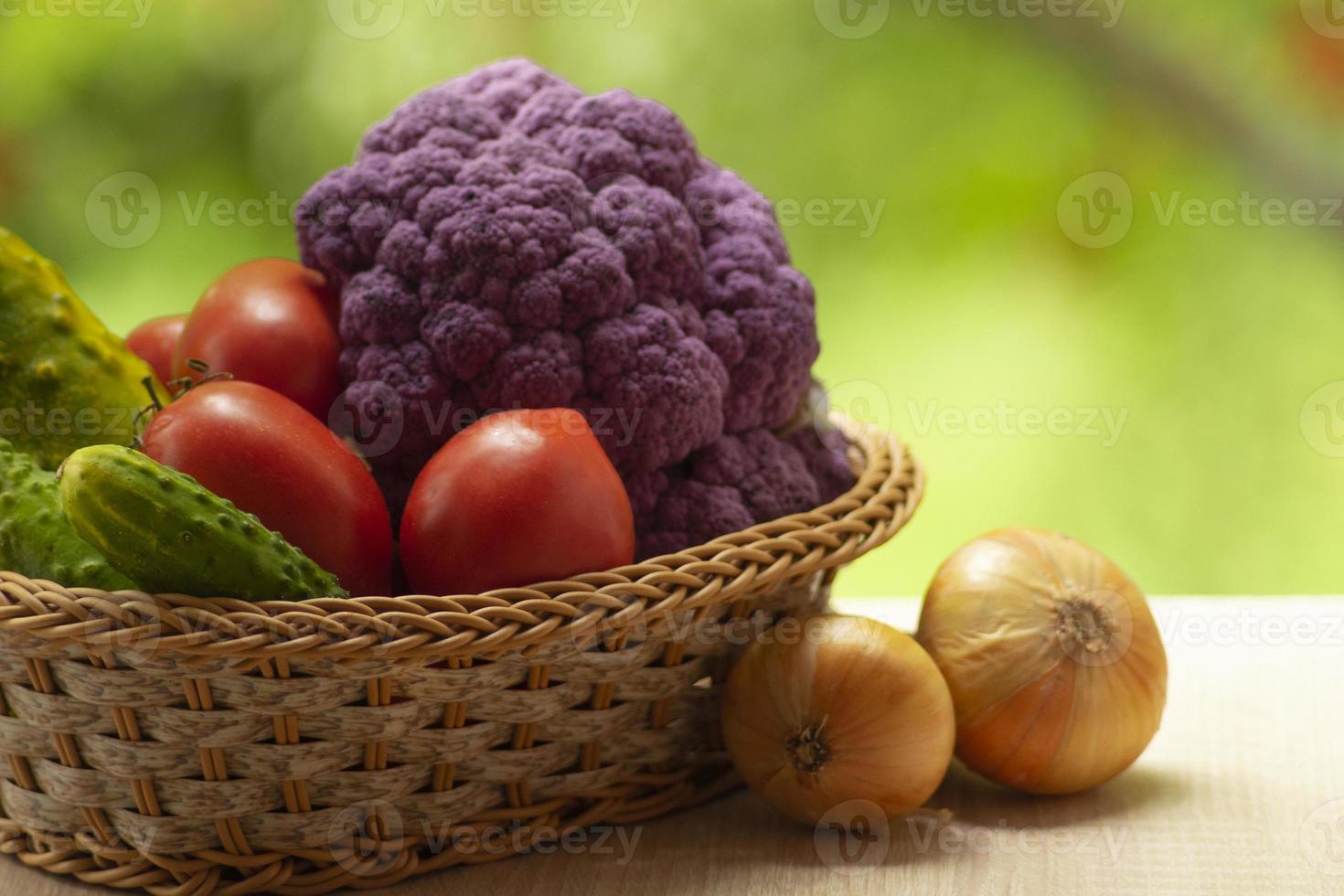 Fresh ripe purple cauliflower, onion, red tomatoes and green cucumber in basket. Healthy food on table on defocus autumn background. photo