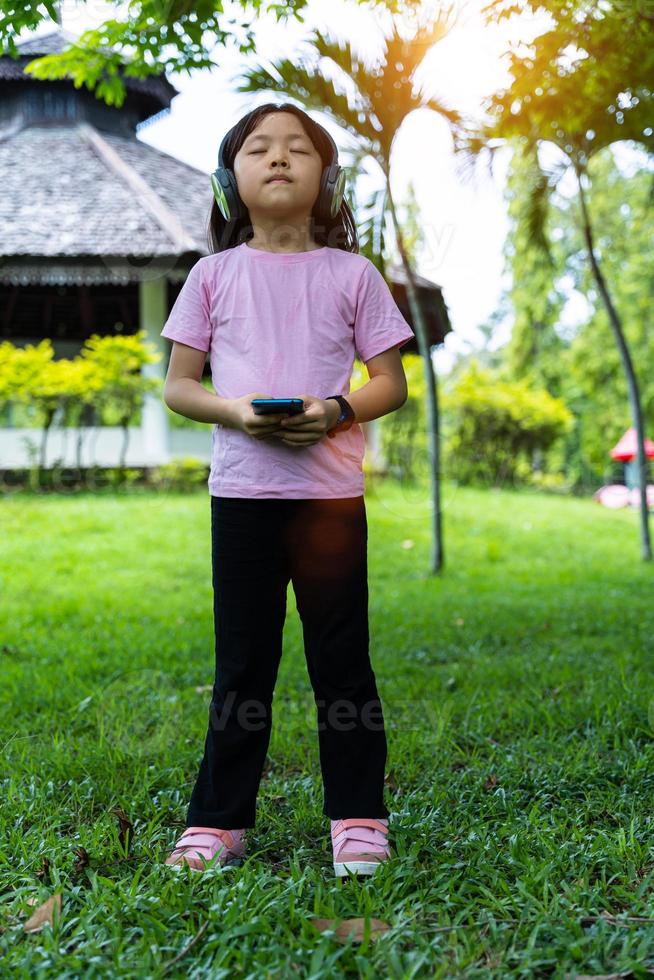 Portrait of child girl with smartphone and listens to music with modern headphones wireless in park outdoors. photo