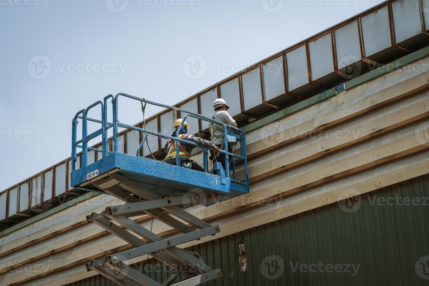 Two workers man on Scissor hydraulic lift or X-lift and use Electric wheel grinding or angle grinder on factory roof at a construction site, Mobile aerial work platform photo