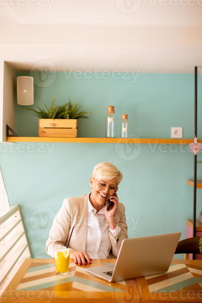 Senior woman using mobile phone while working on laptop and drinking fresh orange juice in the cafe photo