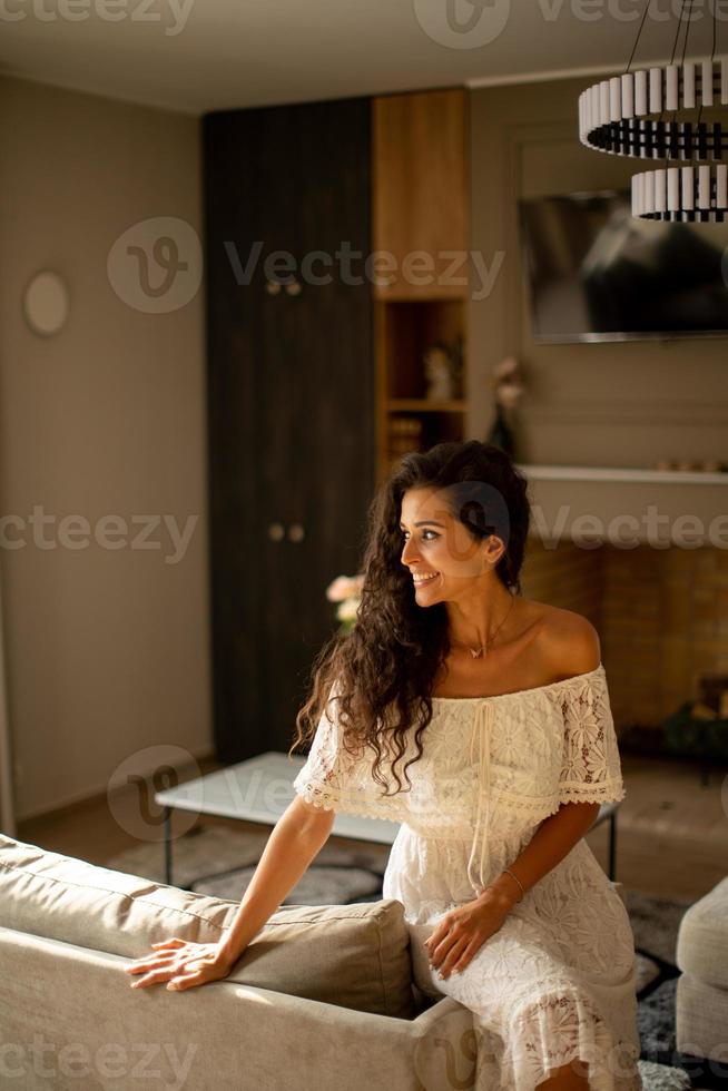 A young woman with curly hair is relaxing in the comfort of her own room, dressed in a white flowing dress photo