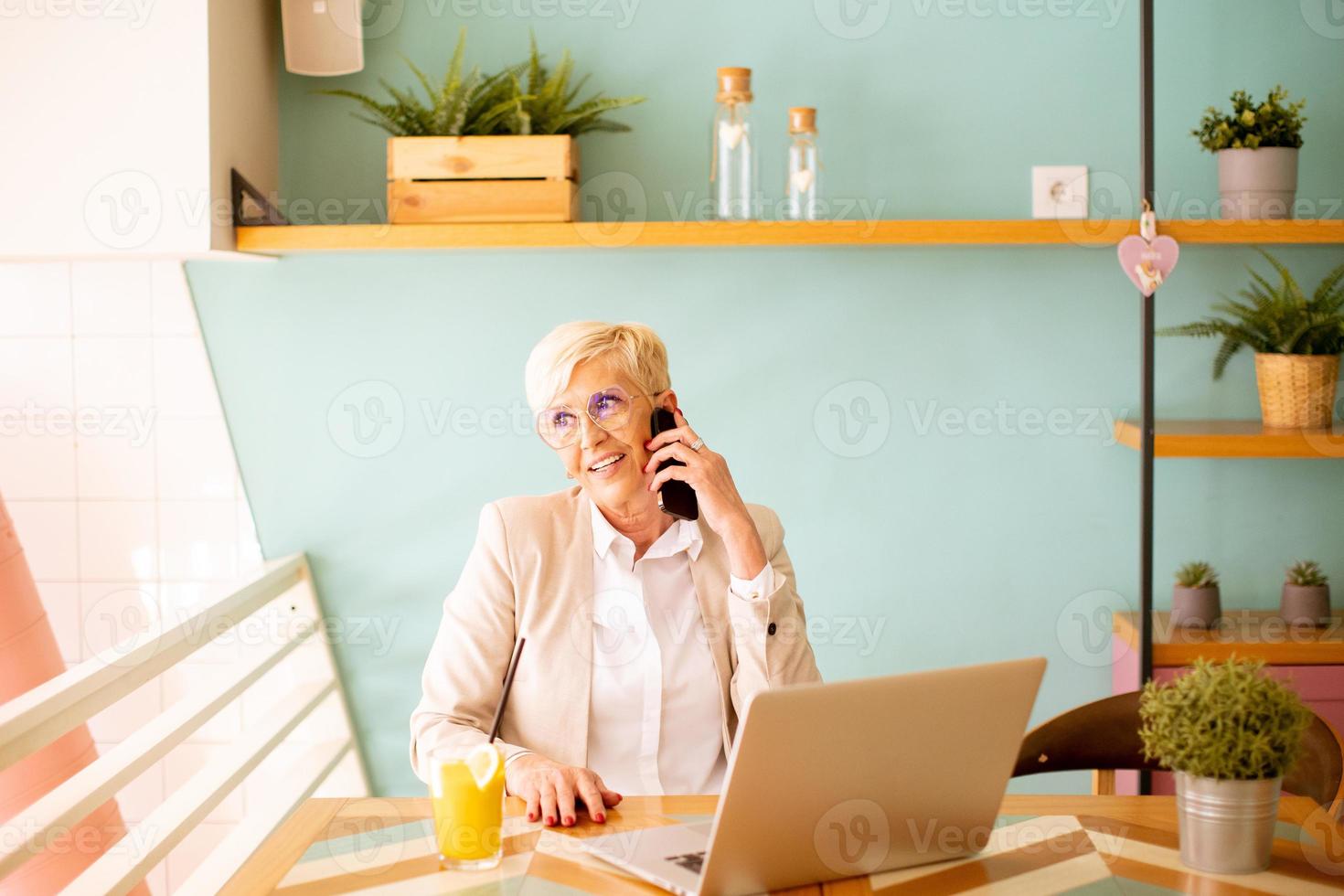 Senior woman using mobile phone while working on laptop and drinking fresh orange juice in the cafe photo