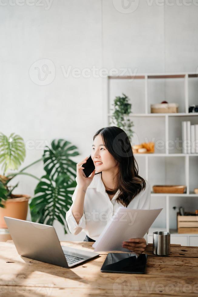 negocio asiático mujer hablando en el teléfono y utilizando un ordenador portátil con un sonrisa mientras sentado a hogar oficina foto