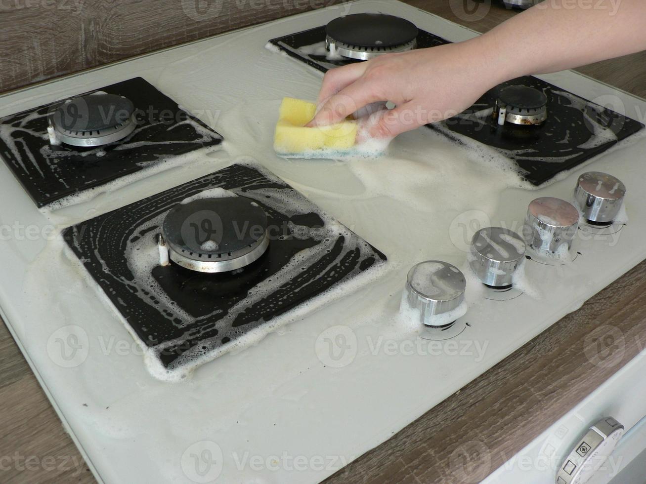 Cleaning dirty gas stove, foam from household chemicals. Hands hold a washing sponge in the foam. White glass hob surface. The concept of household chores, house cleaning. photo