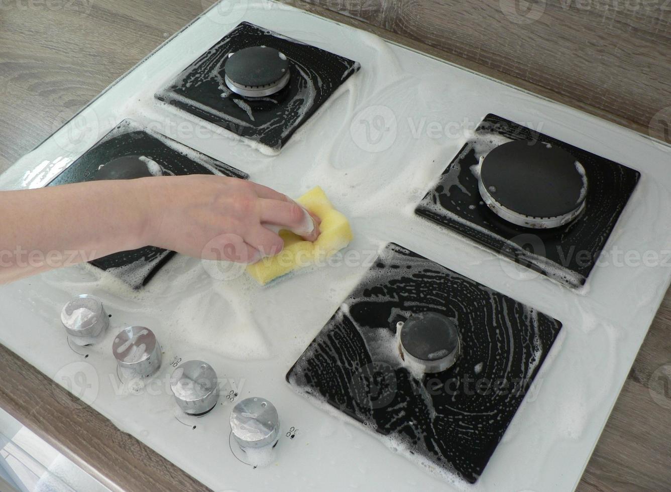 Cleaning dirty gas stove, foam from household chemicals. Hands hold a washing sponge in the foam. White glass hob surface. The concept of household chores, house cleaning. photo