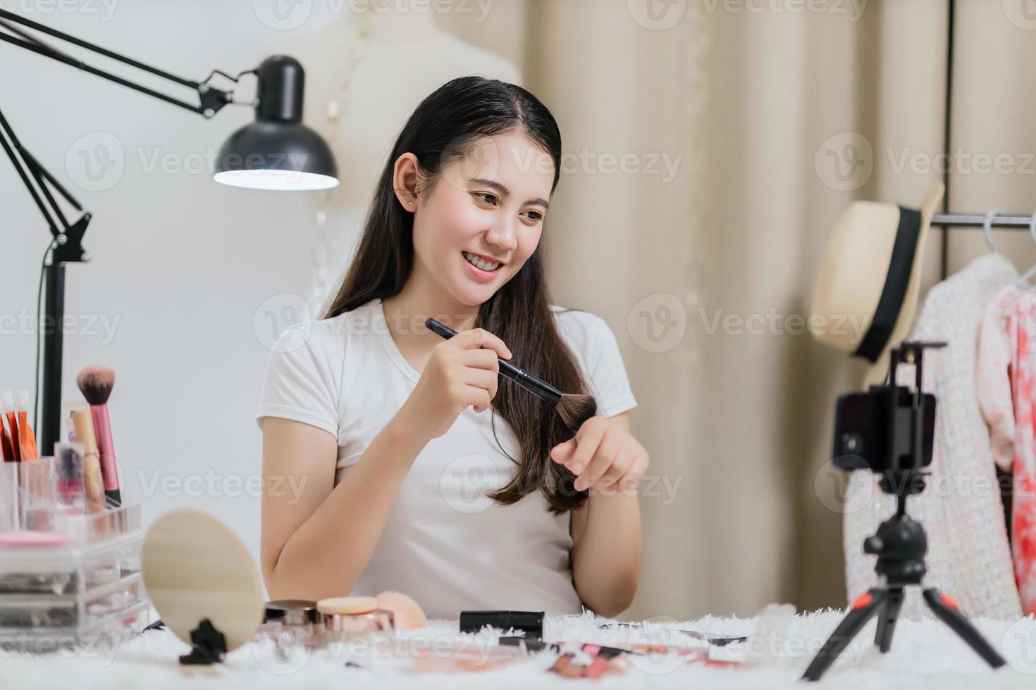 Beautiful Asian woman blogger showing how to make up and use cosmetics live streaming online to social media at home with a smartphone. Beauty blogger or influencer online concept. photo