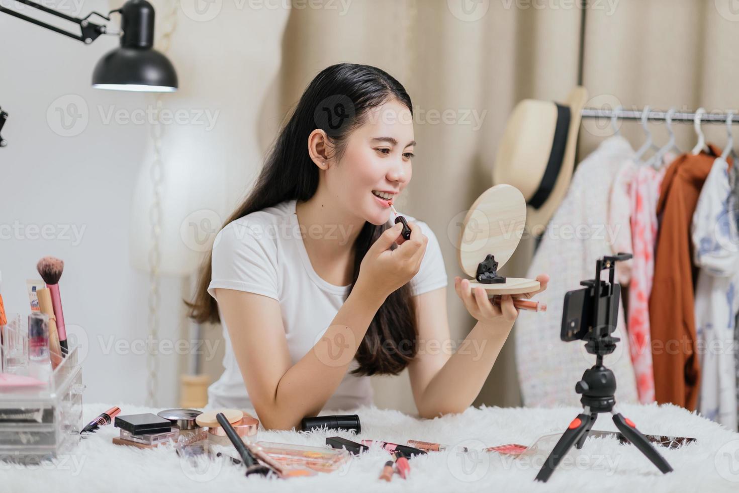 Beautiful Asian woman blogger showing how to make up and use cosmetics live streaming online to social media at home with a smartphone. Beauty blogger or influencer online concept. photo