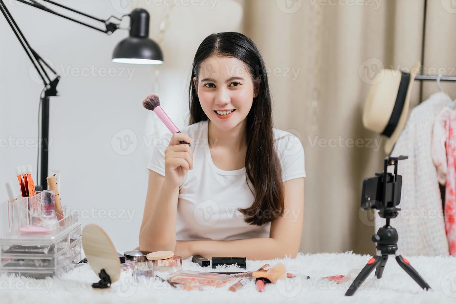 Beautiful Asian woman blogger showing how to make up and use cosmetics live streaming online to social media at home with a smartphone. Beauty blogger or influencer online concept. photo