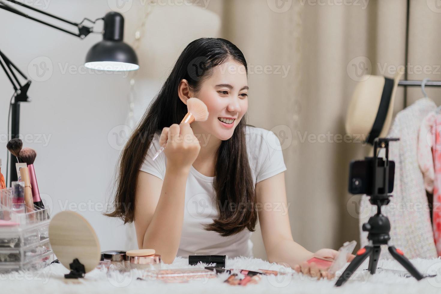 Beautiful Asian woman blogger showing how to make up and use cosmetics live streaming online to social media at home with a smartphone. Beauty blogger or influencer online concept. photo