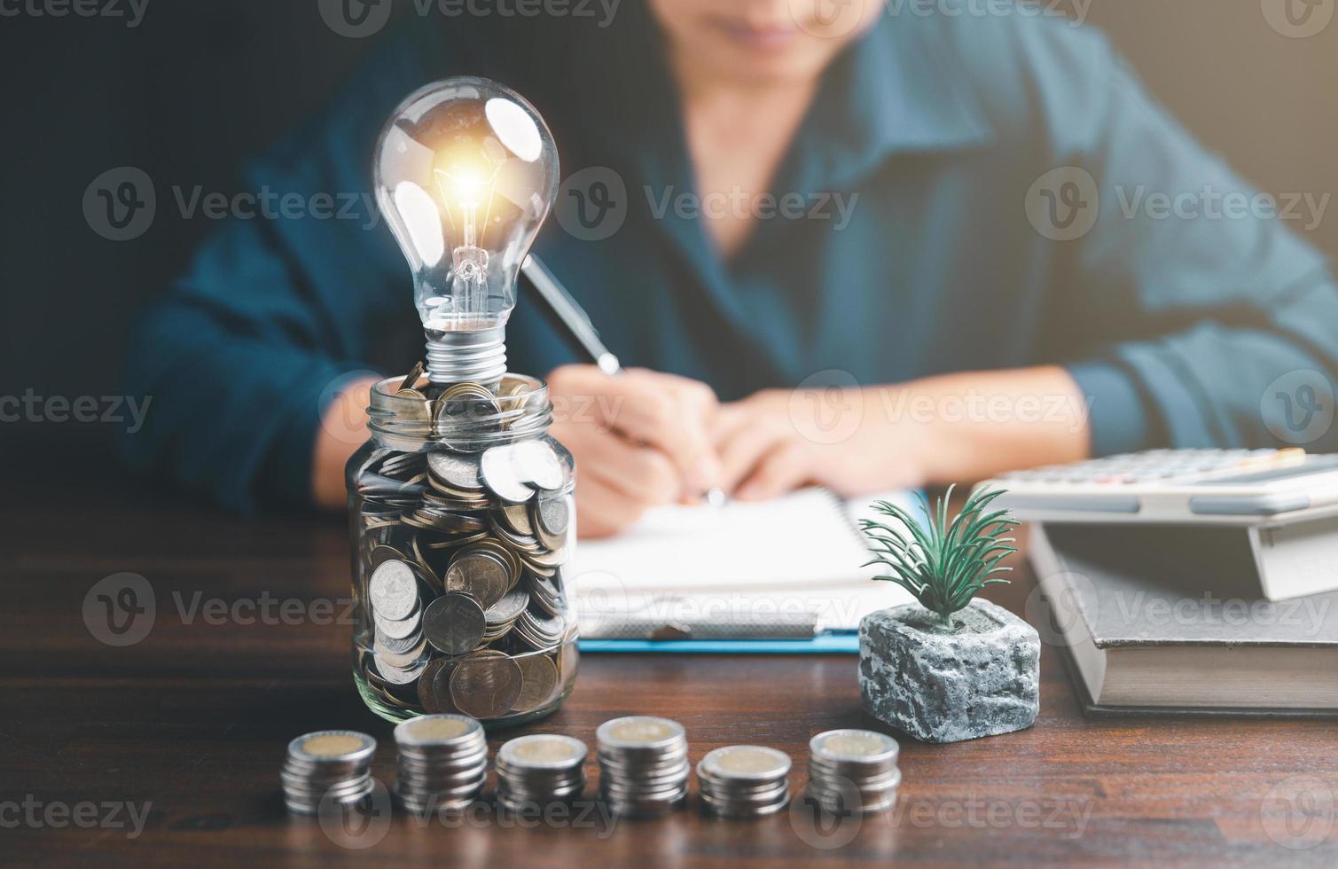 Businesswoman with lightbulb on money jar and using calculator to calculate and money stack. Save energy and money with accounting finance in office concept. Idea of energy saving planning in home. photo