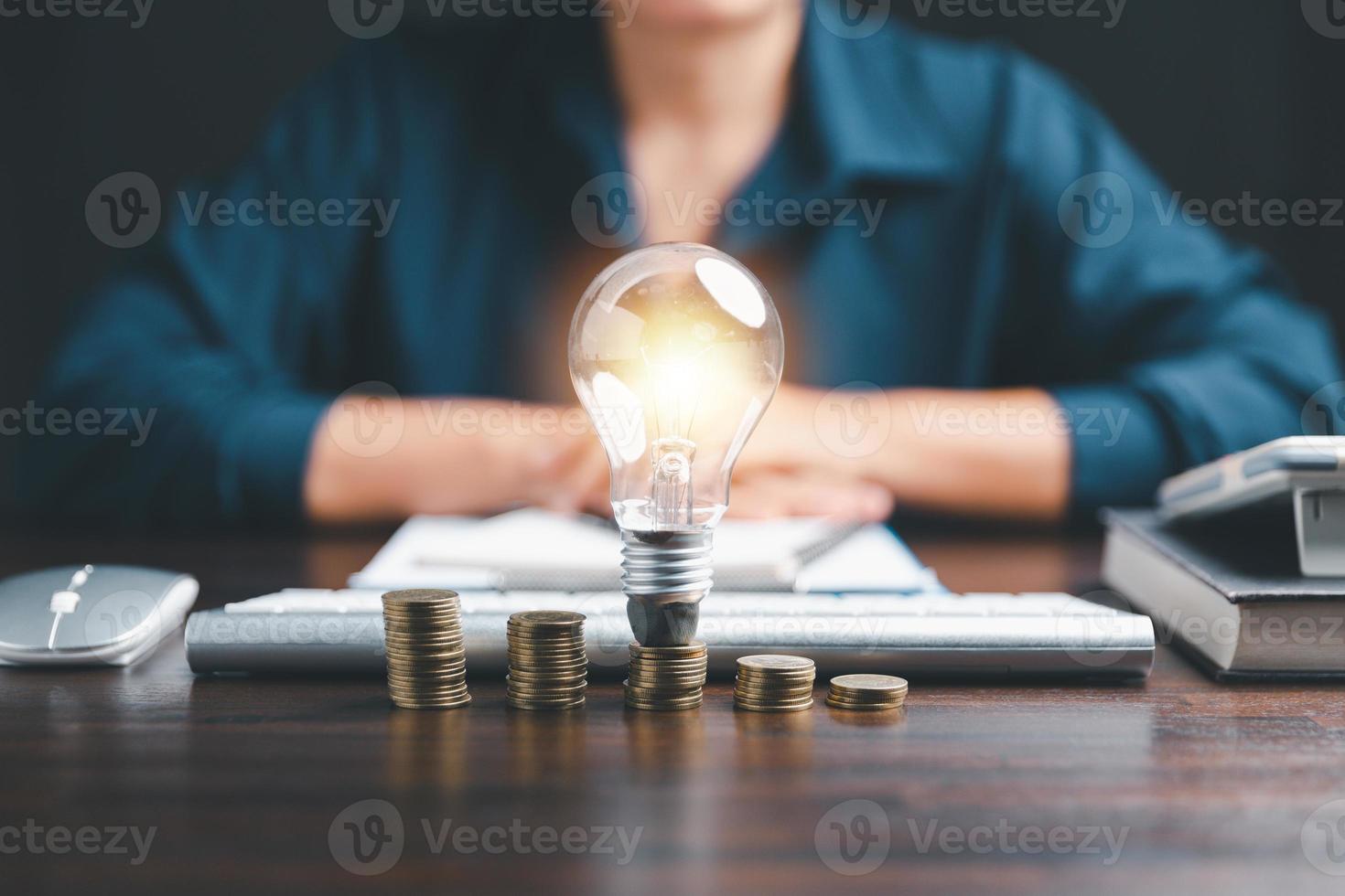Businesswoman with lightbulb on coins and using calculator to calculate and money stack. Save energy and money with accounting finance in office concept. Idea of energy saving planning in home. photo