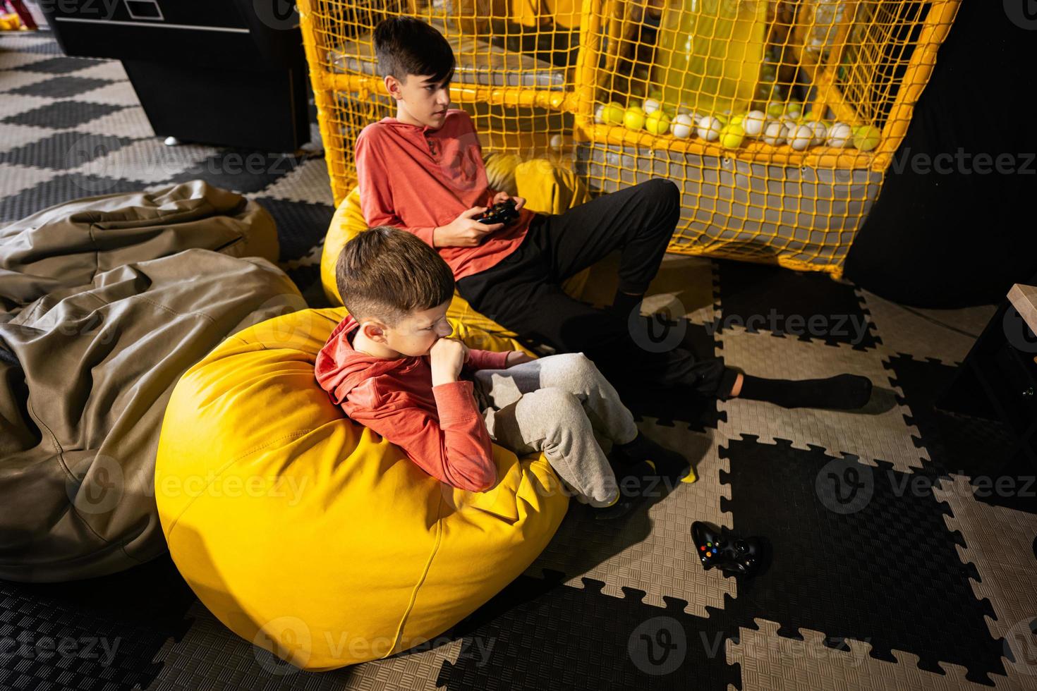 Two brothers playing video game console, sitting on yellow pouf in kids play center. photo