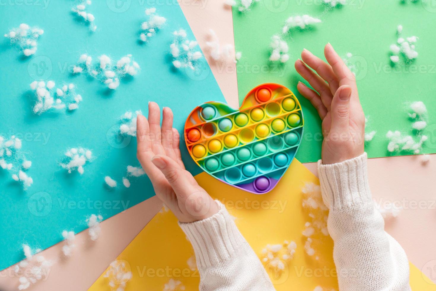 Female hands surround a silicone rainbow antistress toy in the shape of a heart on a cardboard of different colors and fluff. Trendy relaxation tool photo