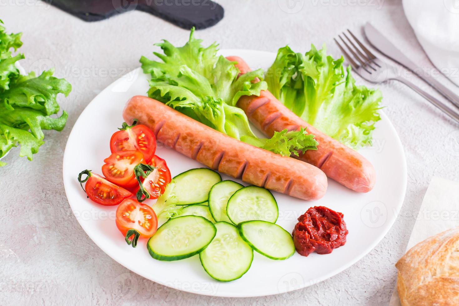 Listo a comer frito salchichas, Cortado Tomates y Pepino vegetales en un plato y un bollo en el mesa. abundante hecho en casa bocadillo foto