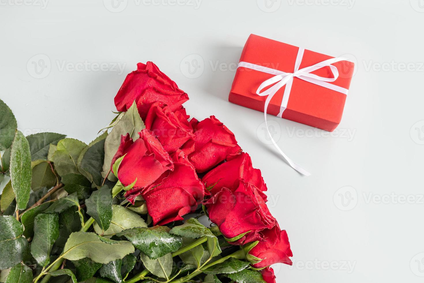 un grande hermosa ramo de flores de rojo rosas con gotas de agua en un ligero antecedentes y un festivo rojo caja con un blanco cinta. parte superior vista. regalo concepto. foto