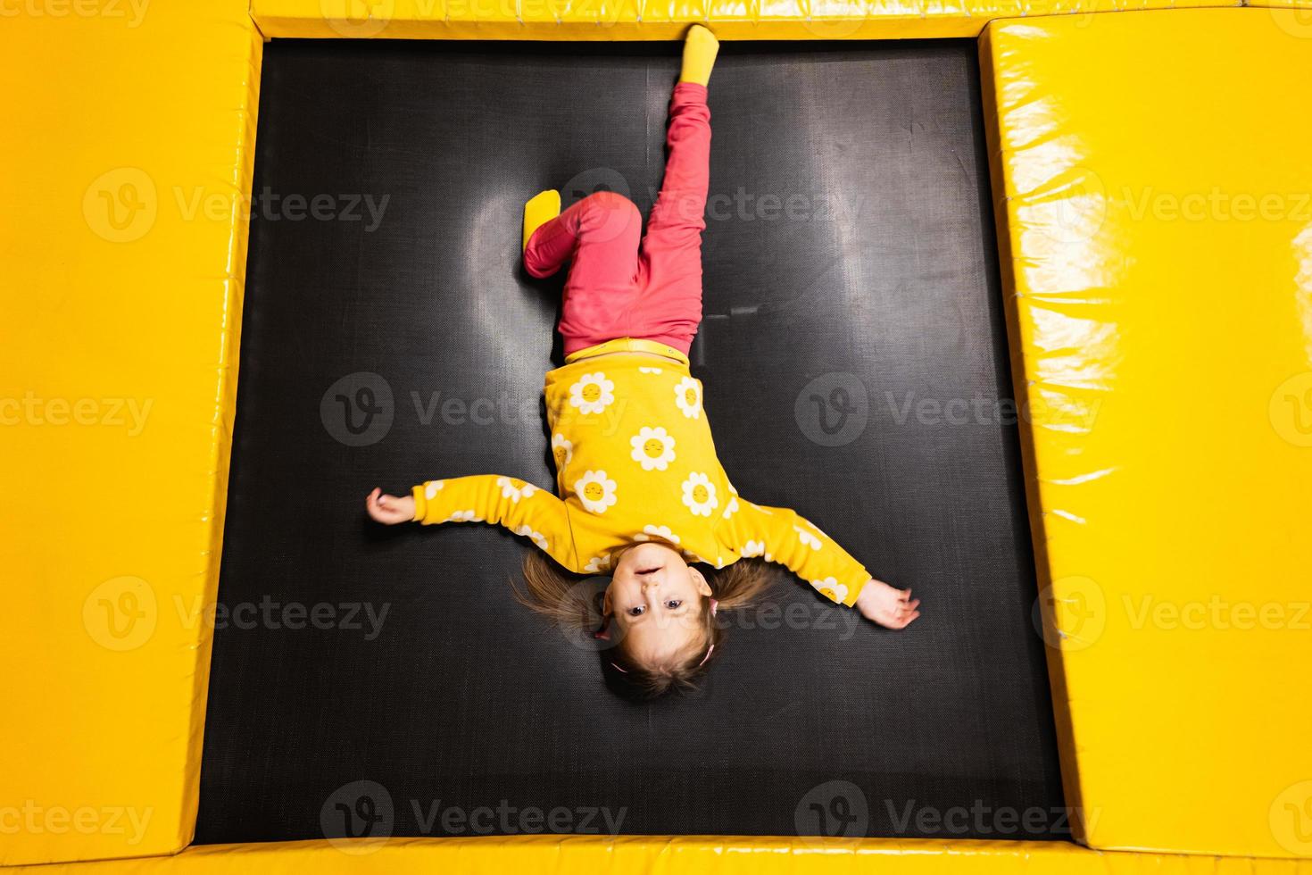 Baby girl kid lies upside down on trampoline at playground park. Child in motion during active entertaiments. photo