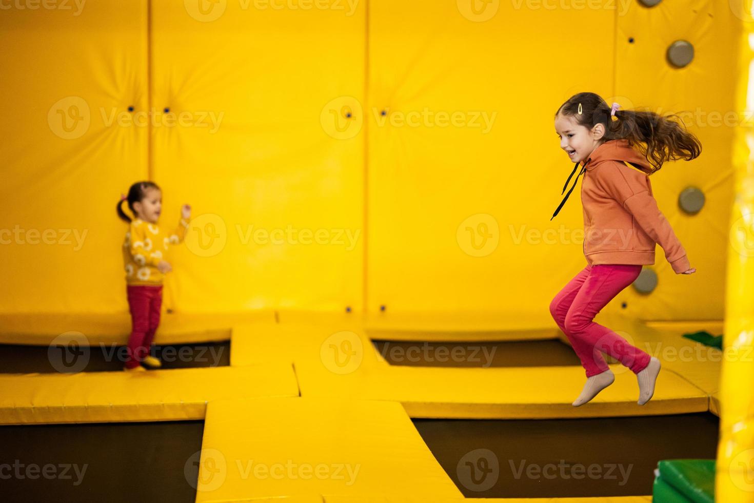 Siblings jumping on trampoline at yellow playground park. Sisters in motion during active entertaiments. photo
