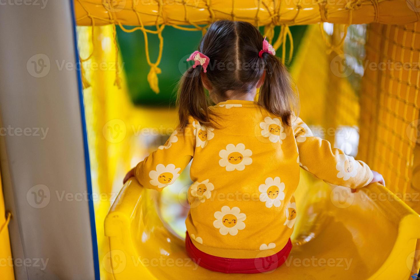 Back of baby girl kid slide at yellow playground park. Child in active entertaiments. photo