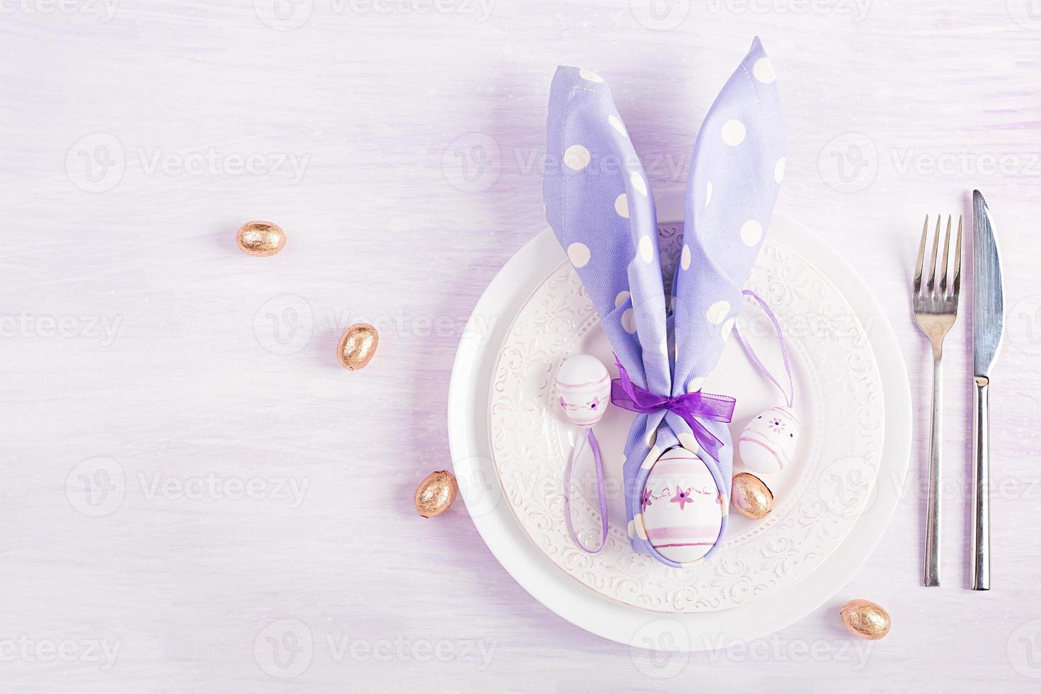 Easter table setting. White plate with a napkin folded in the shape of a rabbit, Easter and chocolate eggs on a pink background. Happy Easter holiday concept. Top view, flat lay photo