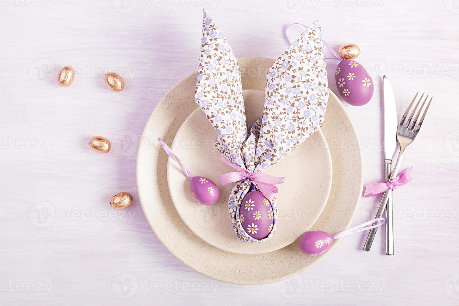 Easter table setting. White plate with a napkin folded in the shape of a rabbit, Easter and chocolate eggs on a pink background. Happy Easter holiday concept. Top view, flat lay photo