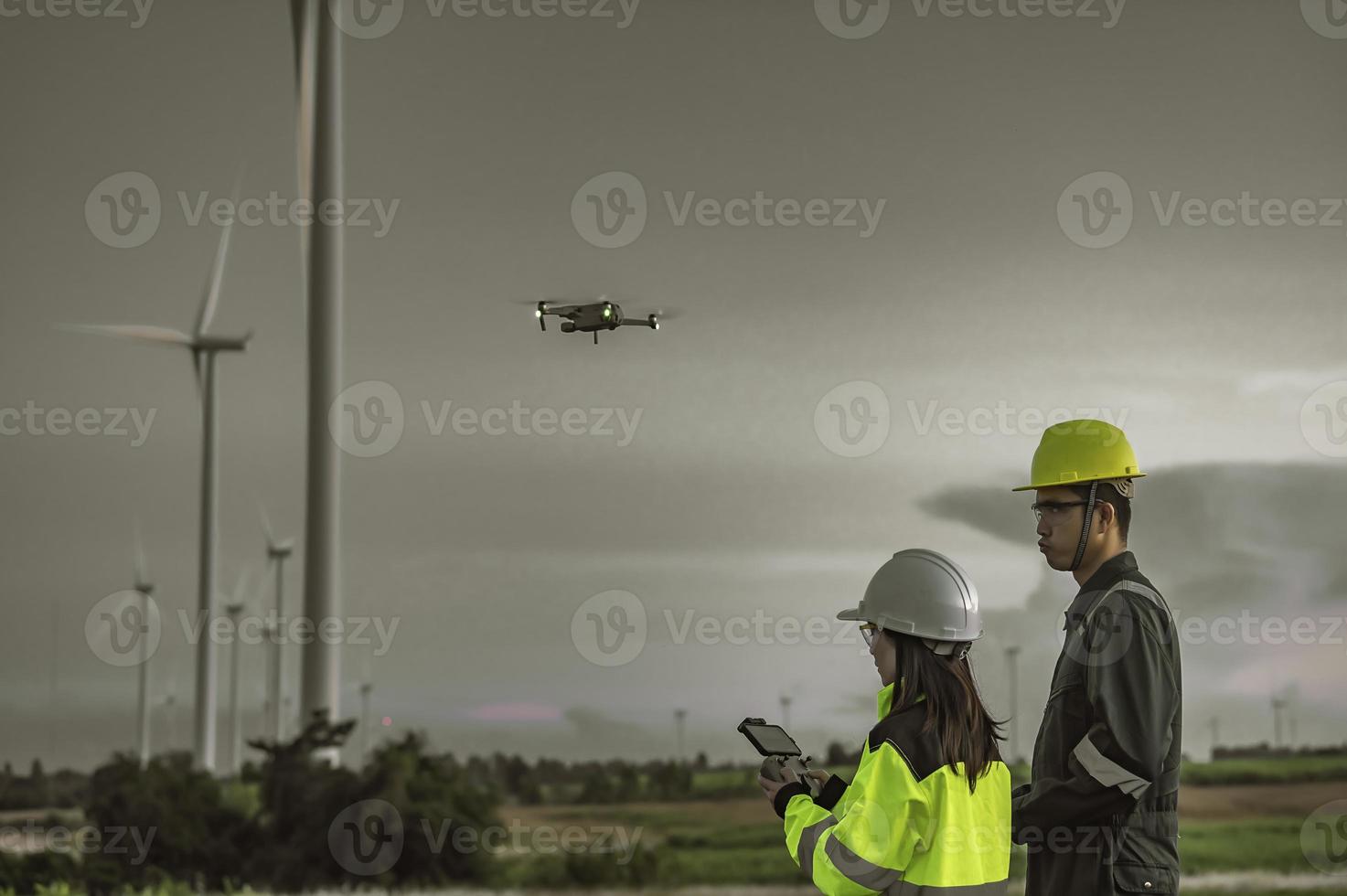 Two engineers working and holding the report at wind turbine farm Power Generator Station on mountain,Thailand people,Technician man and woman discuss about work ,Use drone view from high angle photo