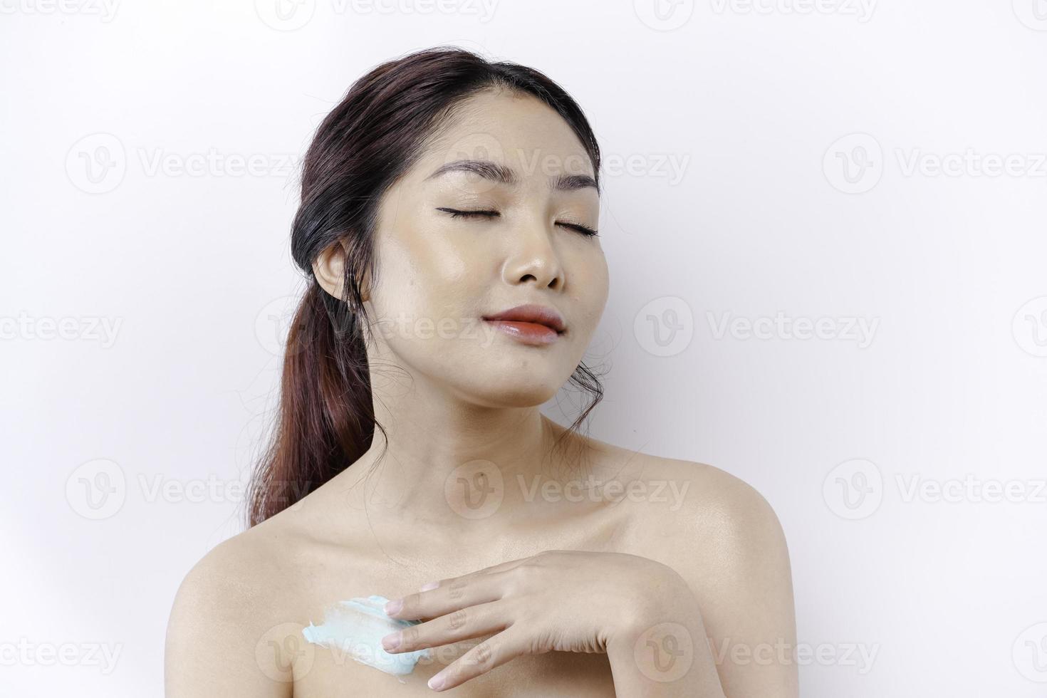 piel cuidado productos concepto. asiático mujer aplicando hidratante loción en cuerpo después ducha, en pie envuelto en toalla, recortado imagen foto