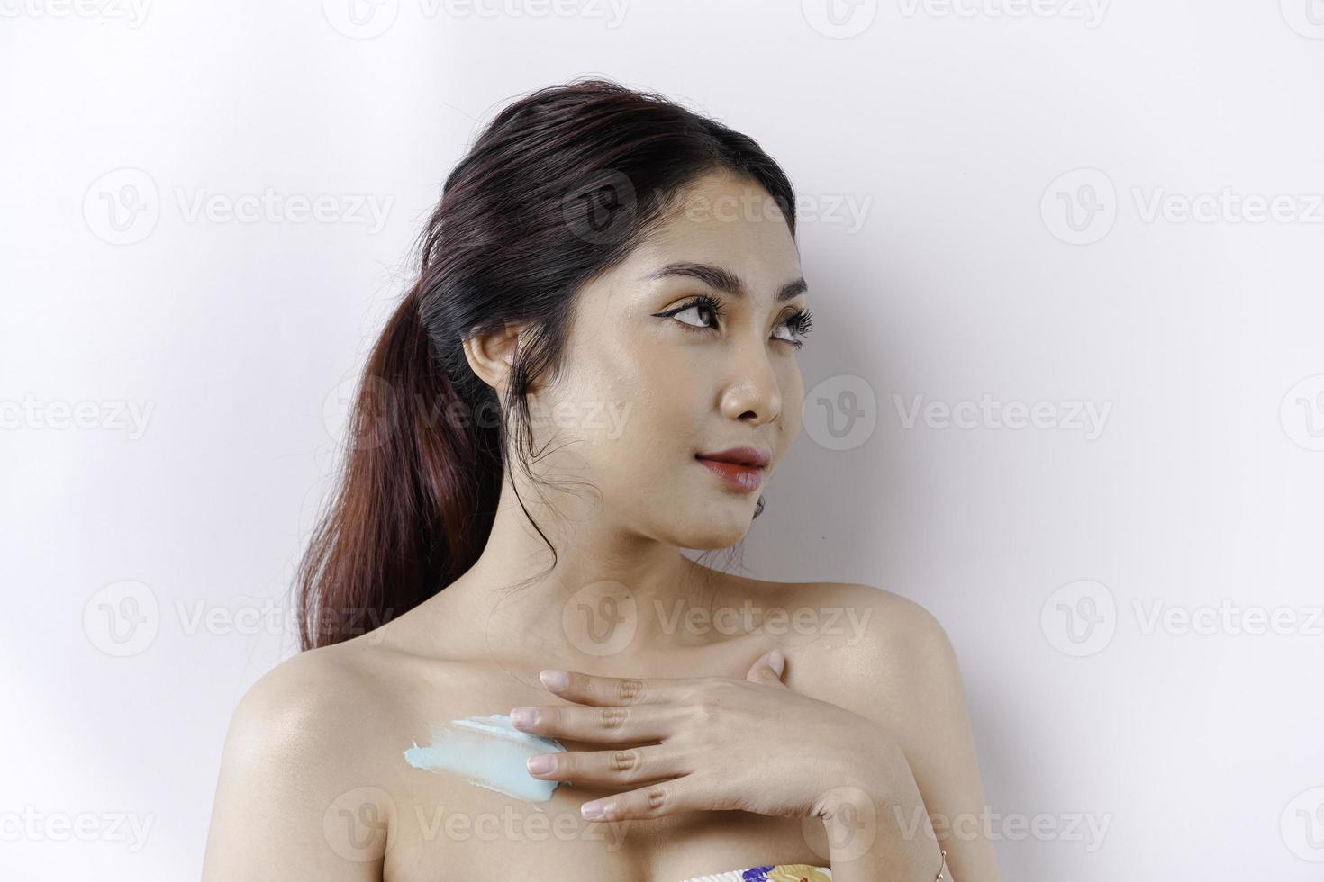 piel cuidado productos concepto. asiático mujer aplicando hidratante loción en cuerpo después ducha, en pie envuelto en toalla, recortado imagen foto