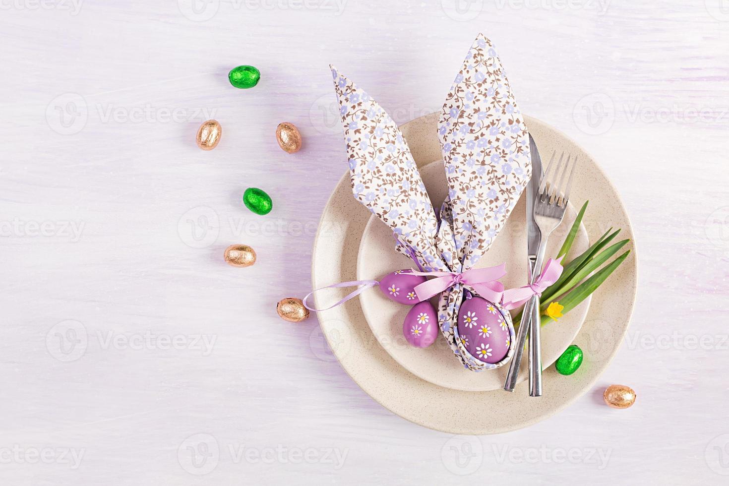 Easter table setting. White plate with a napkin folded in the shape of a rabbit, Easter and chocolate eggs on a pink background. Happy Easter holiday concept. Top view, flat lay photo