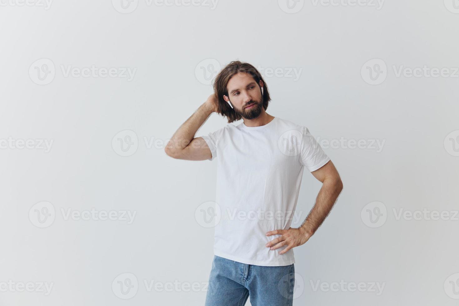 A man with a beard and long hair in a white T-shirt and blue jeans stands against a white wall, leaning against it and listening to music with wireless white headphones, staring thoughtfully photo