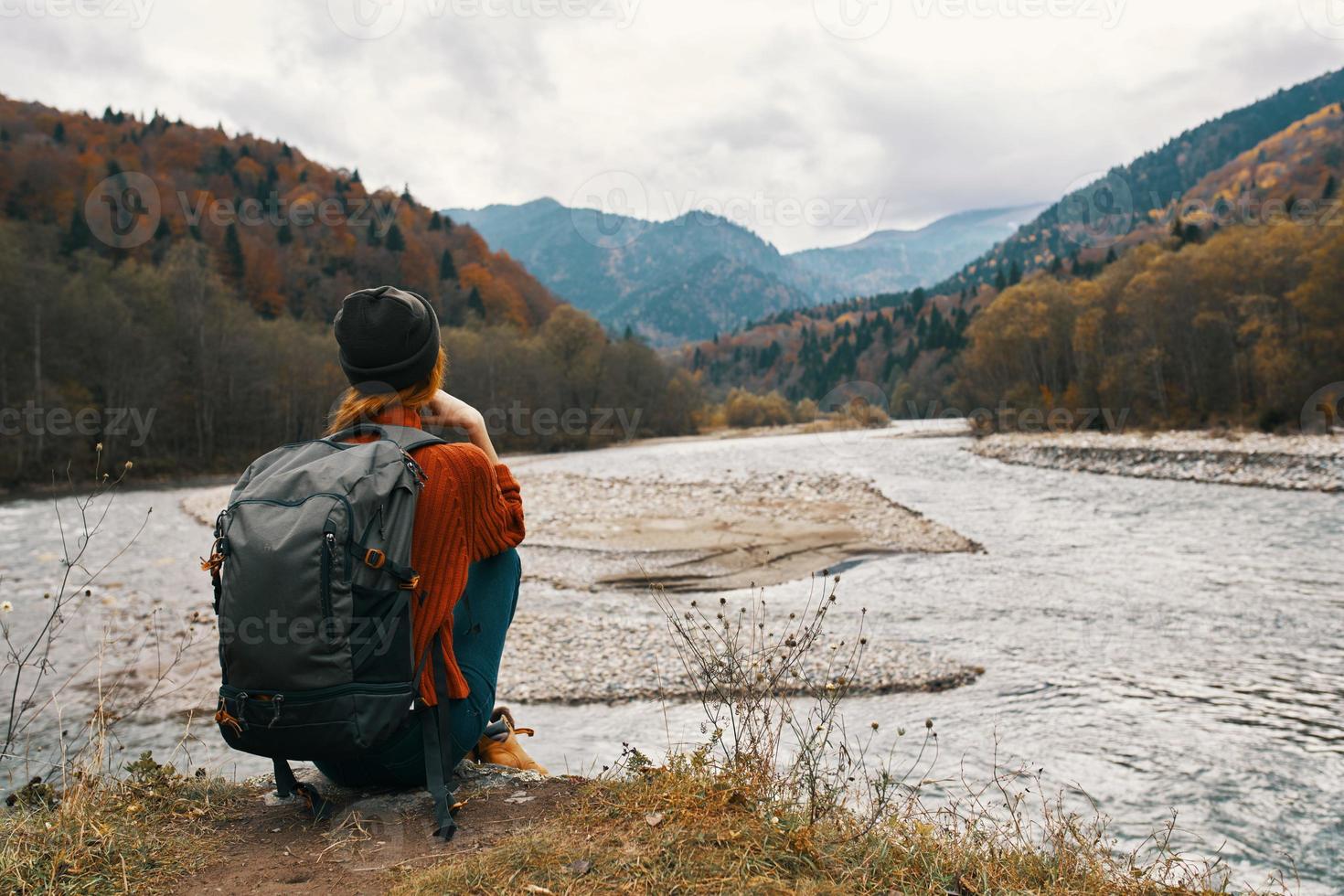 woman in the mountains in autumn with a backpack Sits on the river bank and looks at the high mountains photo