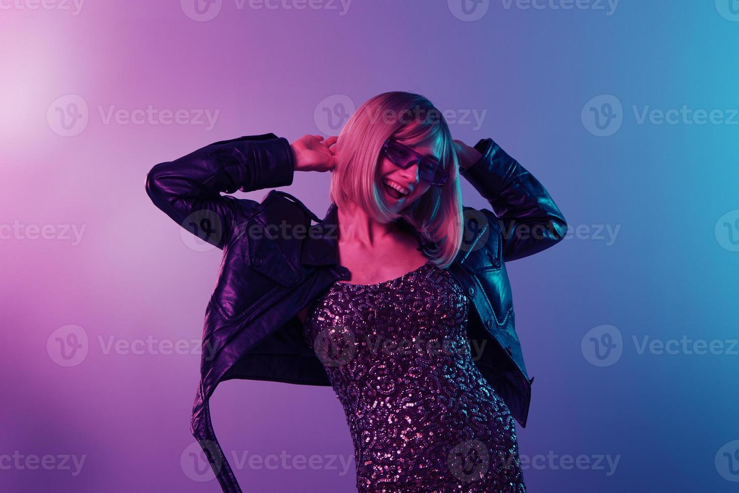 Sexy beautiful blonde lady in leather jacket sparkly dress trendy sunglasses dance hold hands up posing isolated in blue pink color light studio background. Neon party Cyberpunk concept. Copy space photo