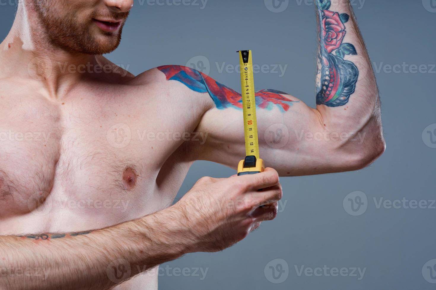 guy measures arm muscles with centimeters on a gray background and a multicolored tattoo bodybuilder fitness photo