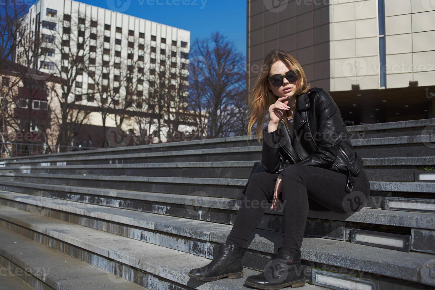 Woman in leather jacket sits on the stairs near the building in the autumn on the street photo