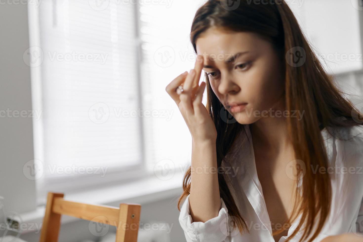woman with eyeliner sitting at a table indoors, window in the background photo