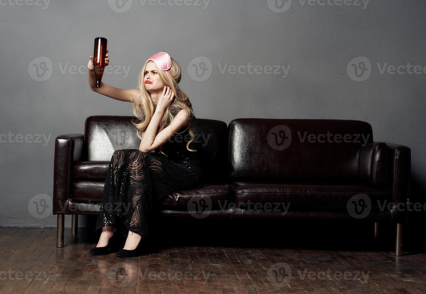 A blonde woman with a bottle of beer on a leather sofa in a black dress photo