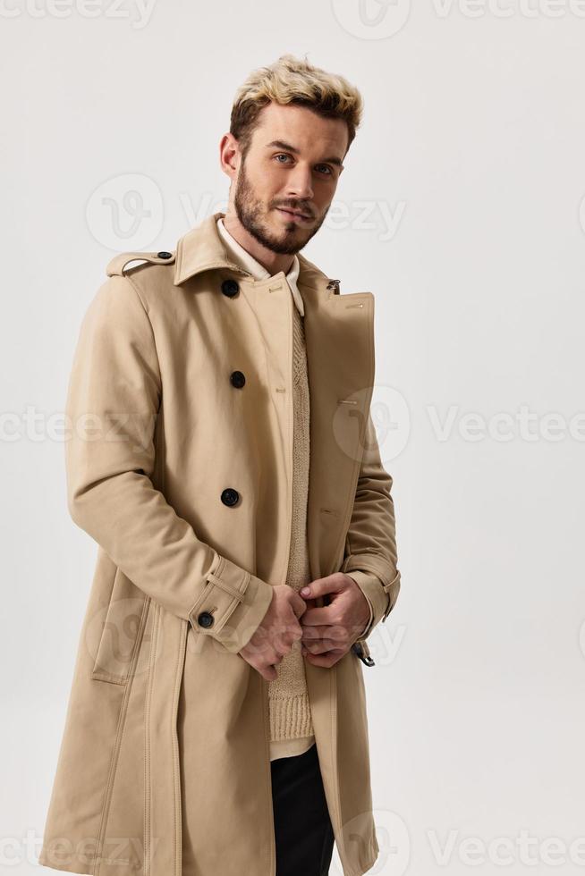 handsome blond man in a beige coat holds his hands in his pockets on a light background photo