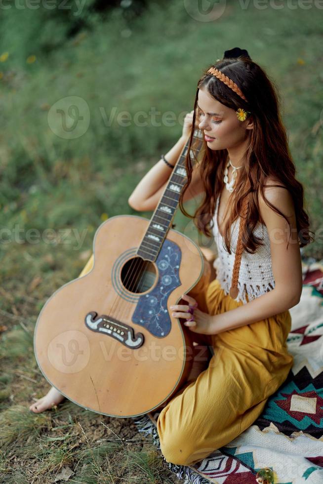 Hippie woman eco friendly playing guitar smiles and singing songs in nature sitting on a plaid by the lake in the evening in the sunset sunlight. A lifestyle in harmony with the body and nature photo