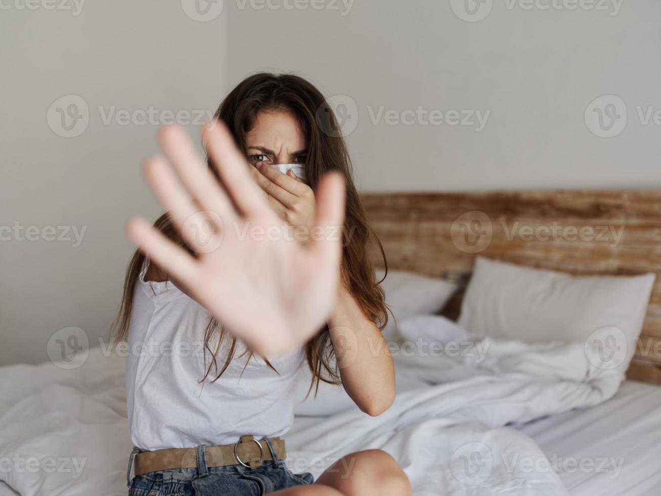 woman in medical mask covers her face with hand and sits on bed indoors photo