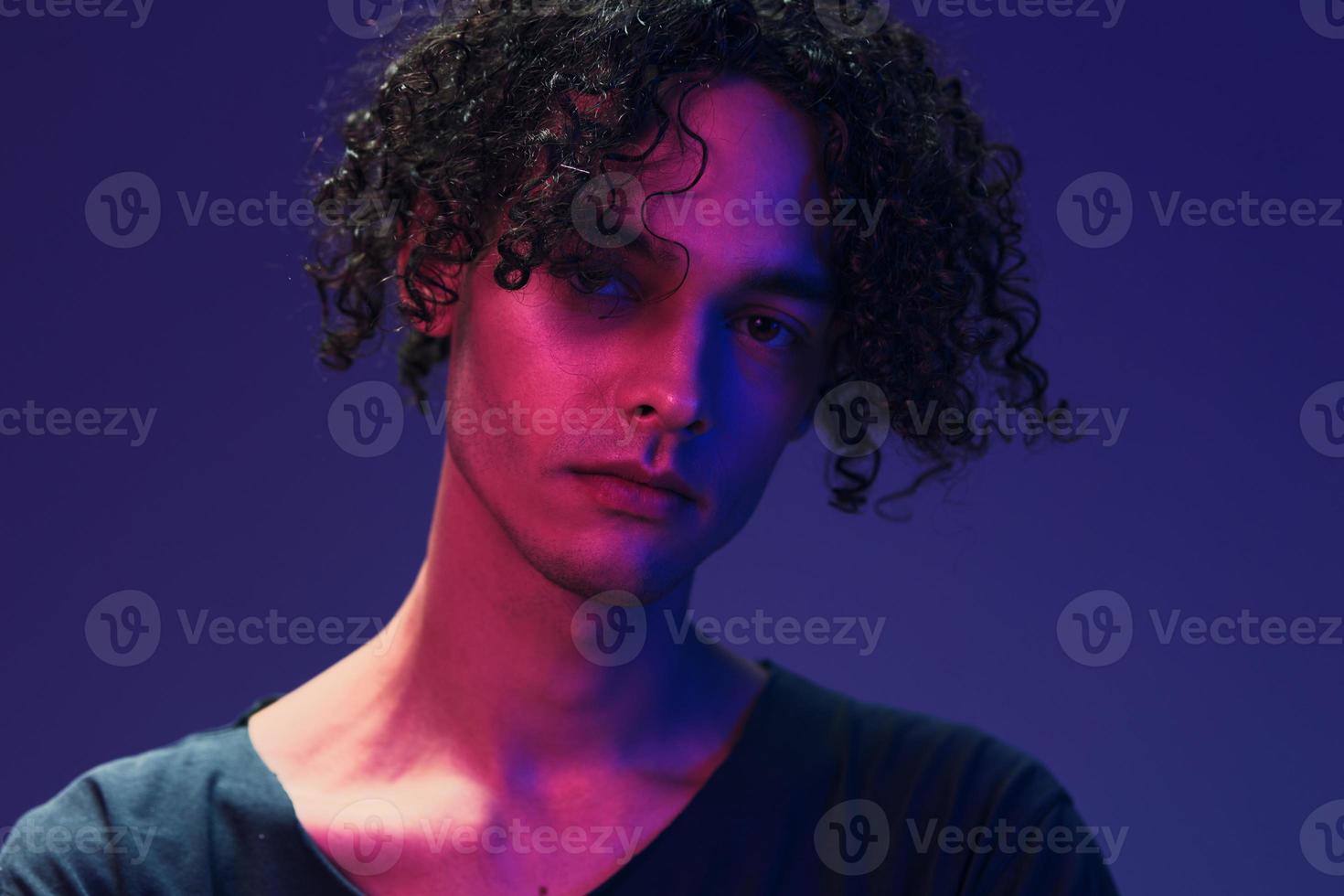 Stylish tanned curly man in black t-shirt looks at camera posing isolated on color pink blue studio background. Cool fashion offer. Virtual Reality New Collection concept. Copy space for ad photo