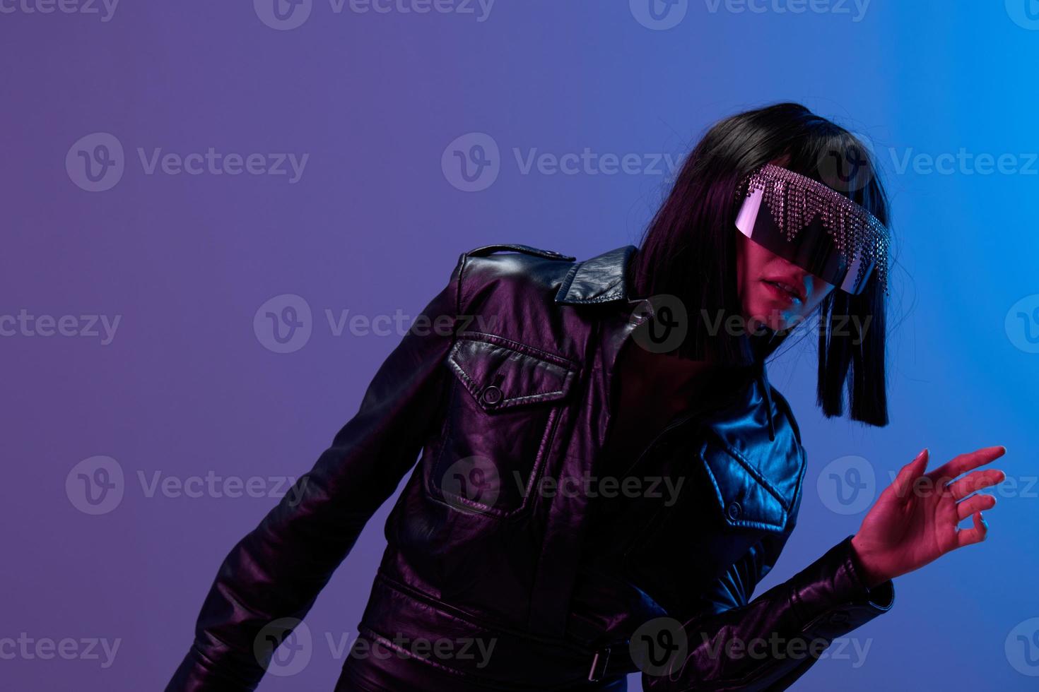 Confused adorable brunet woman in leather jacket specular sunglasses open mouth look aside posing isolated in blue violet color light background. Neon party Cyberpunk concept. Copy space. Good offer photo