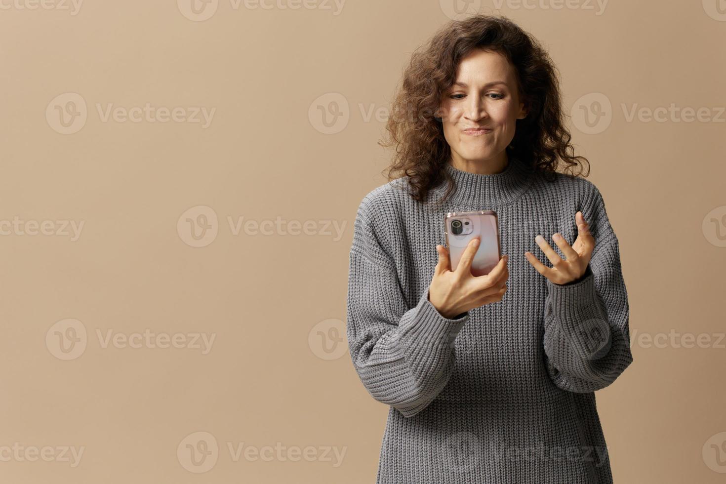 Evil irritated curly beautiful female in gray casual sweater squeeze phone with fury posing isolated on over beige pastel background. Social media, network, distance communication concept. Copy space photo