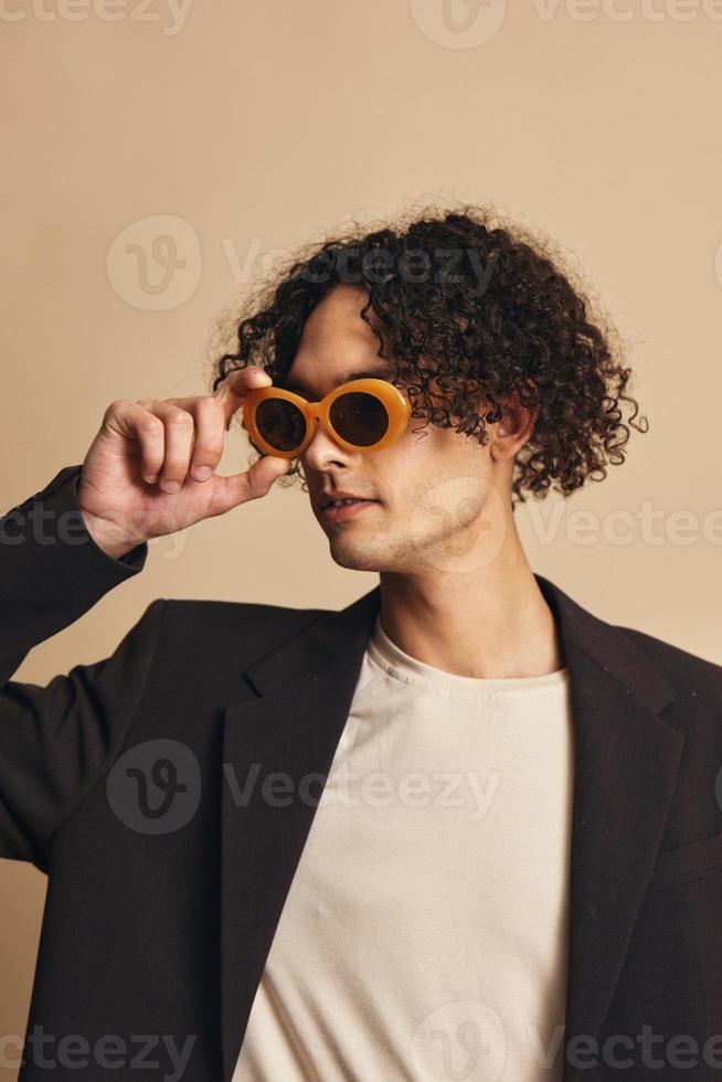 Demonstrative awesome tanned curly man in classic brown jacket trendy sunglasses posing isolated on over beige pastel background. Fashion New Collection offer. Retro style concept. Free place for adDemonstrative awesome tanned curly man in classic brown j photo