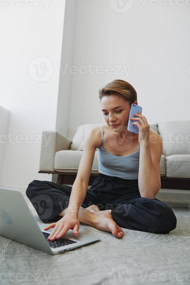 Freelance woman with laptop and phone works from home sitting on the floor in her home clothes with a short haircut, free copy space photo
