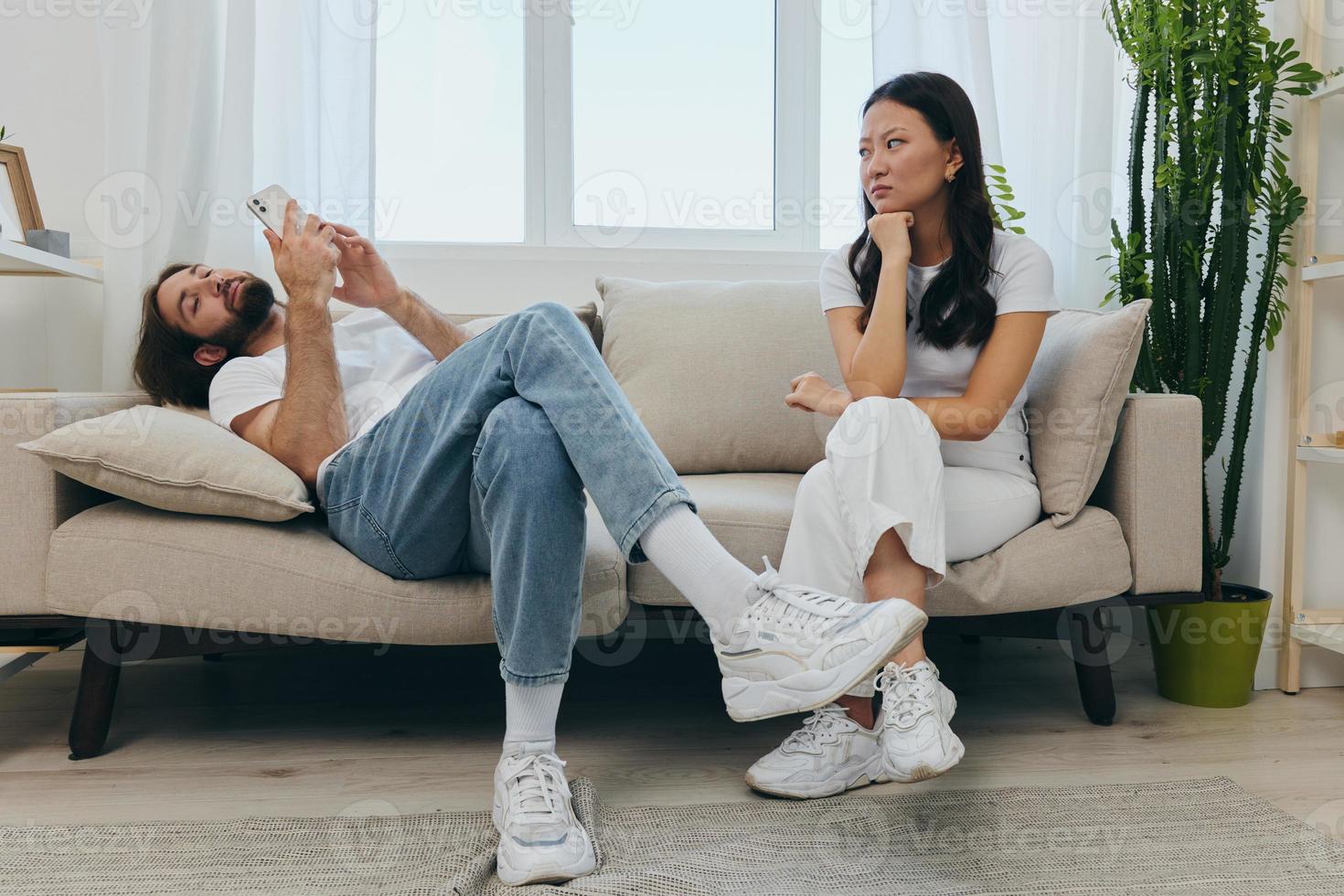 A man looks at his phone screen and smiles during an argument with his girlfriend. The angry and hurt woman looks in his direction and is sad. Family discord at home, phone addiction photo