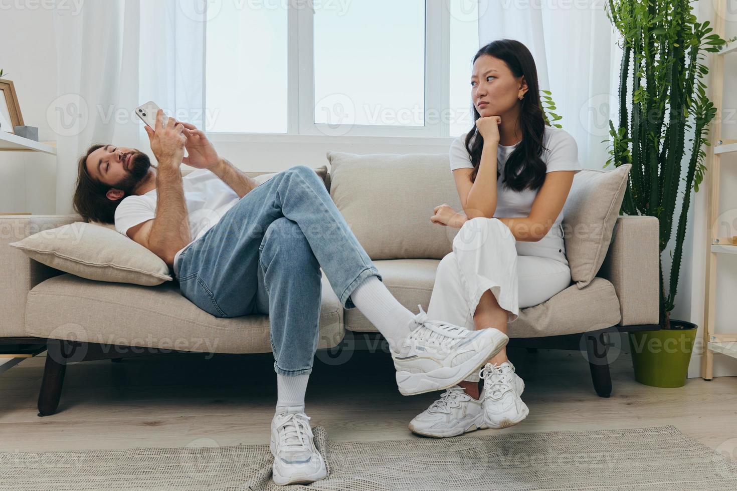 un hombre mira a su teléfono pantalla y sonrisas durante un argumento con su novia. el enojado y herir mujer mira en su dirección y es triste. familia discordia a hogar, teléfono adiccion foto