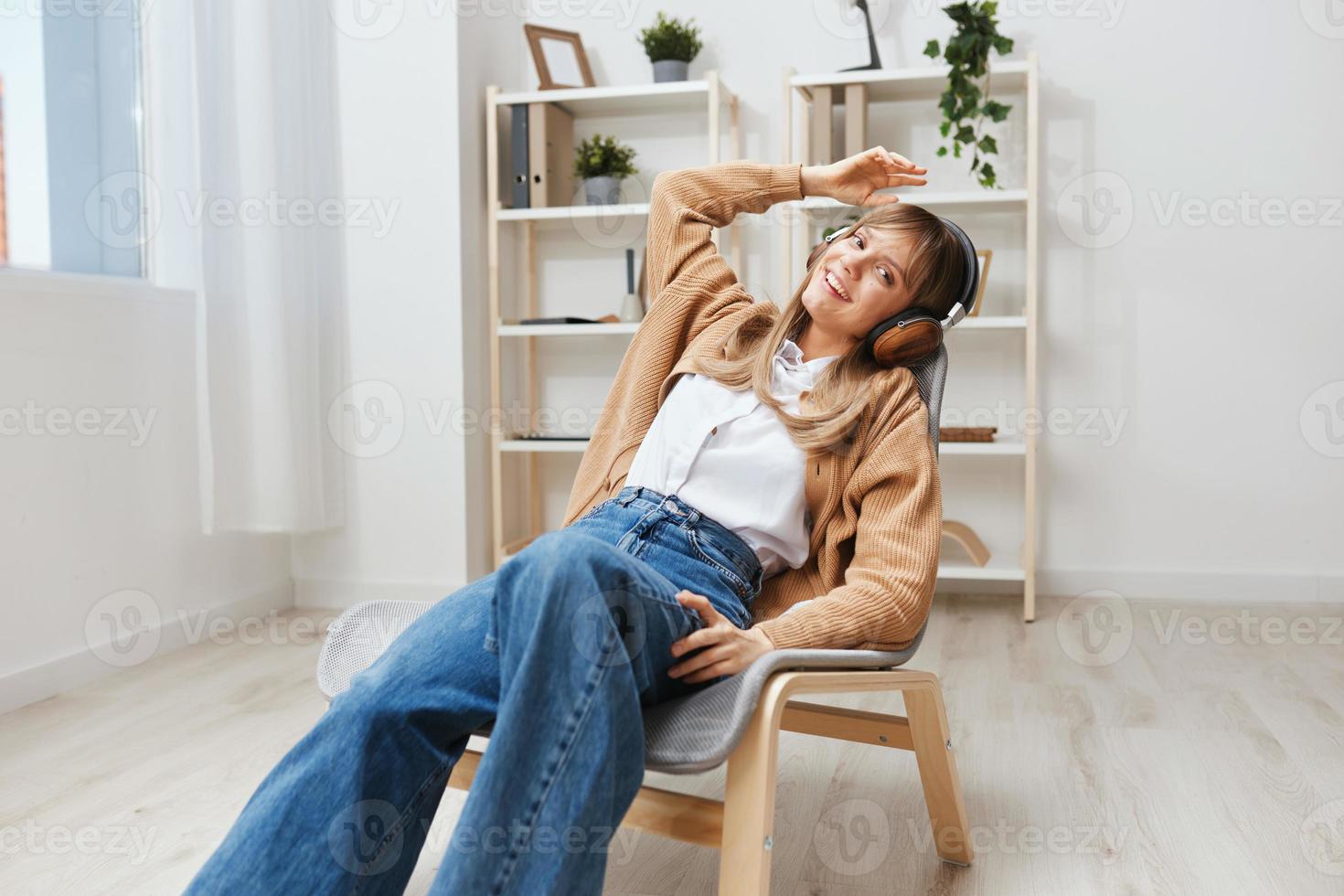 Happy smiling young blonde lady in warm sweater in headphones listen moves to favorite songs sitting in armchair at modern home interior. Music time Relaxing Cool playlist Concept. Copy space photo