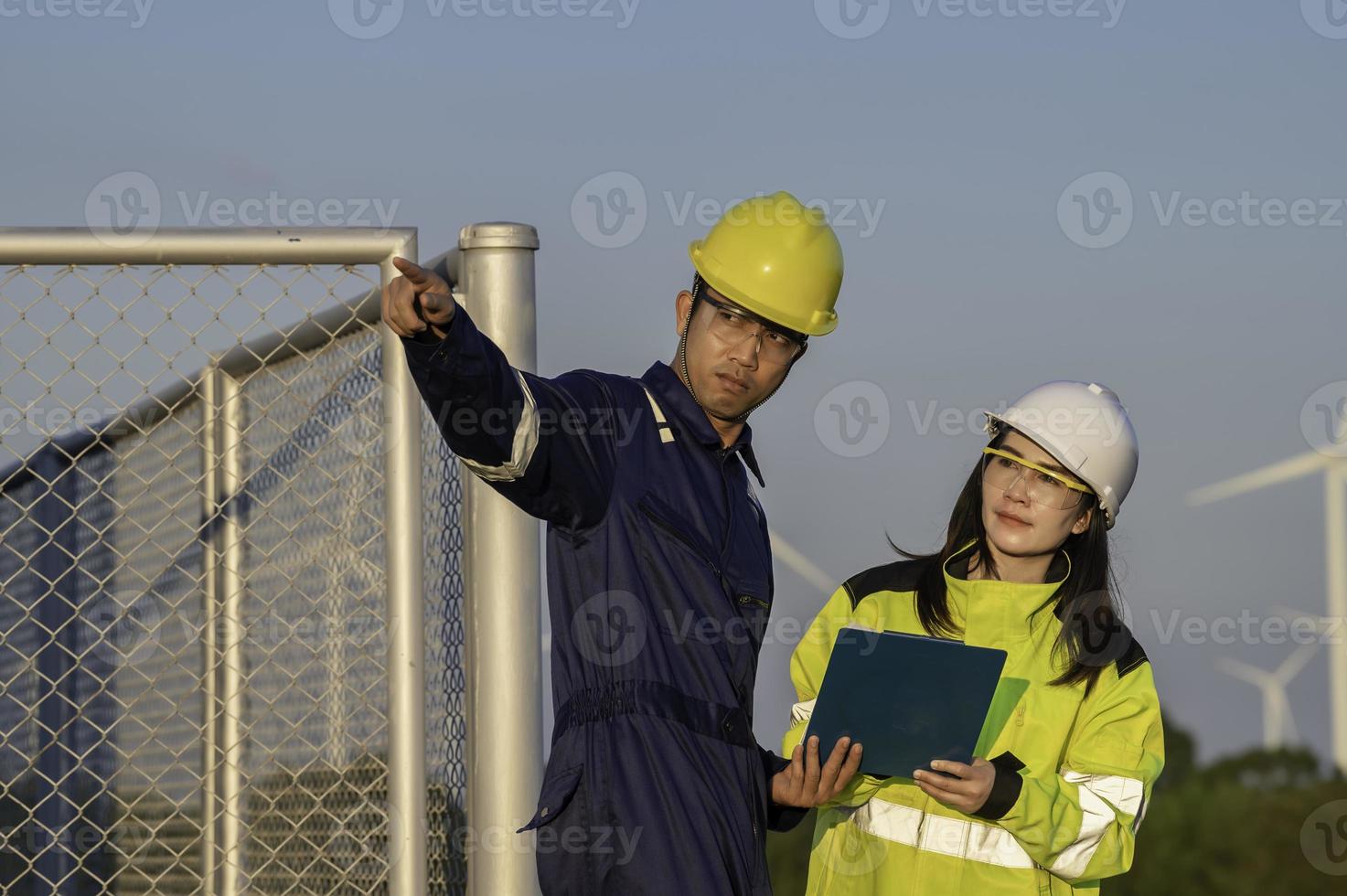 Two engineers working and holding the report at wind turbine farm Power Generator Station on mountain,Thailand people,Technician man and woman discuss about work ,Use drone view from high angle photo