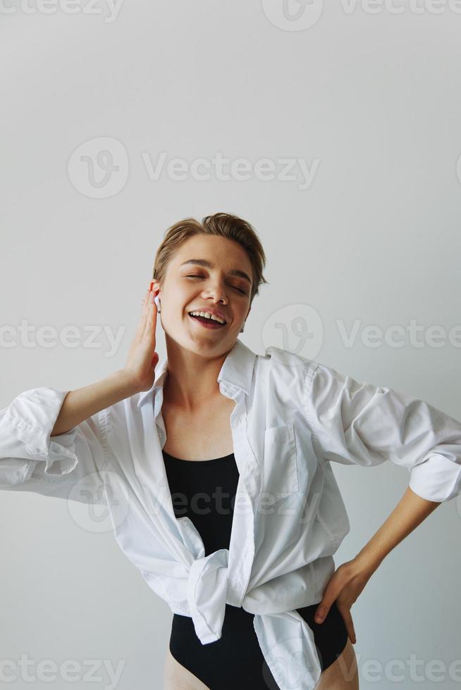 joven mujer adolescente escuchando a música con estéril auriculares y bailando hogar, sonriendo con dientes con un corto Corte de pelo en un blanco camisa en un blanco antecedentes. niña natural poses con No filtros foto